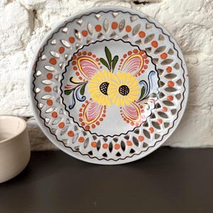Op de afbeelding: Een decoratieve keramische plaat met een grijze achtergrond en een bloemmotief. De plaat heeft een gele bloem met zwarte centra, omgeven door roze, oranje en groene accenten. De rand heeft uitgesneden patronen.
