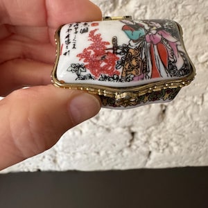 May include: A small, rectangular trinket box with a hinged lid. The box is white with a gold-colored trim and features a colorful illustration of figures in traditional clothing and red flowers. The box is held in a hand.