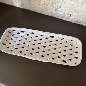 Op de afbeelding: Een rechthoekige witte keramische mand met een geweven ontwerp. De mand heeft een rechthoekige vorm met afgeronde hoeken en een vlechtwerkpatroon. De mand is leeg en staat op een donkere ondergrond.