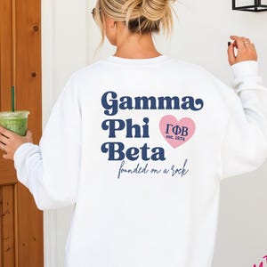 May include: A white crewneck sweatshirt featuring dark blue text on the back that reads Gamma Phi Beta, with a pink heart containing the Greek letters ΓΦΒ and the text est. 1874, followed by the script text founded on a rock below the heart.