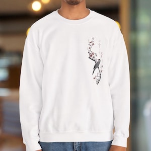 以下が含まれることがあります： 白いスウェットシャツ。日本の伝統的なスタイルで、鳥が赤い花咲く枝に止まっているイラストが描かれています。左胸に日本語の文字が添えられています。クルーネックのデザインです。