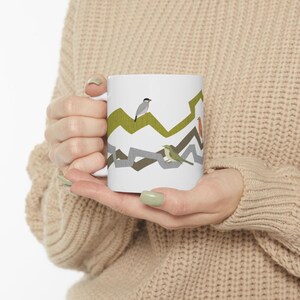 Puede incluir: Taza de cerámica blanca con un diseño de pájaros. El diseño presenta pájaros posados sobre un patrón en zigzag en tonos verdes, grises y marrones. La taza está sujeta por una persona.