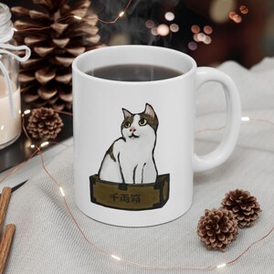 May include: White ceramic mug featuring a cat illustration. The cat, with brown and white markings, is depicted sitting in a box with Japanese text. The mug has a white handle and is surrounded by decorative elements.