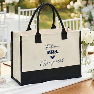 May include: A canvas tote bag with black handles and a black bottom panel. The bag is off-white with the words "Future MRS." and a name printed in navy blue. The bag is on a table with wedding decorations.