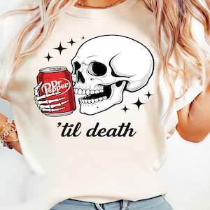 May include: A cream-colored t-shirt featuring a graphic of a white human skull with skeletal hands holding a red Dr. Pepper soda can. The design includes black star accents and the text 'til death written in a black cursive script font below.