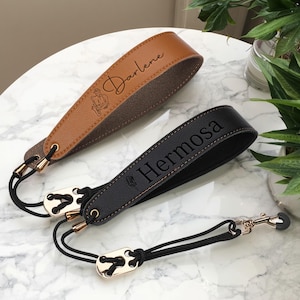 May include: Two personalized leather wrist straps for a pet leash. One is brown with the name "Darlene" and a graphic, the other is black with the name "Hermosa". Both have gold-colored hardware and black rope.