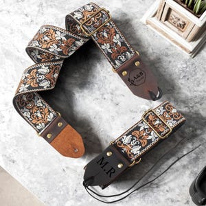 以下が含まれることがあります： 黒、オレンジ、白、シルバーの花柄織りのギター用ストラップ。ストラップは茶色の革の端と金色の金属製の金具が付いています。「A&B」と「M.R.」の刻印がそれぞれ片方の端にあります。