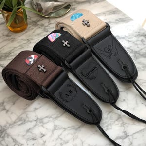May include: Three guitar straps in brown, black, and beige, each with a black leather end piece. The straps feature decorative crosses and personalized engravings, including initials, a heart with a name, and a hand-holding design. Guitar picks are visible.