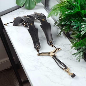 May include: Black leather suspenders with gold-colored hardware. The suspenders have a unique design with multiple adjustment points and the text "RAYMOND CARTER" printed on the leather. The suspenders are displayed on a white marble surface.