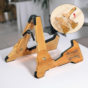 以下が含まれることがあります： パーソナライズされたデザインの木製譜面台。スタンドは、黒のアクセントと金属製の金具が付いた明るい色の木材で作られています。ベースには、「ALEYNA & BERKAY」と書かれた花柄とテキストの詳細があります。