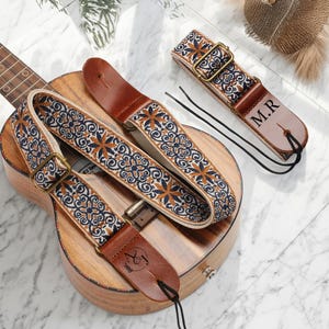 Könnte beinhalten: Eine Ukulele mit zwei dekorativen Gitarrengurten. Die Gurte haben einen cremefarbenen Hintergrund mit einem sich wiederholenden Muster aus marineblauen und orangefarbenen floralen Designs. Die Gurte haben braune Lederenden und Metallschnallen. Ein Gurt hat die Initialen "M.R."