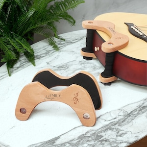 以下が含まれることがあります： 黒いパッドと「Emily Johnson」の名前が刻まれた木製のギターレスト。 1 つのレストはギターに取り付けられ、もう 1 つは大理石の表面に置かれています。 ト音記号も刻まれています。