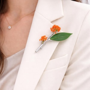 Könnte beinhalten: Silberfarbene Brosche in Form eines Zweigs, verziert mit leuchtend orangefarbenen Blüten und einem einzelnen grünen Blatt. Die Brosche ist an einem weißen Blazer befestigt und bildet einen auffälligen Kontrast.