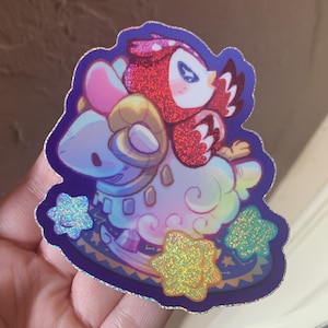 May include: A holographic sticker featuring a red owl perched on a light blue ram. The design includes three sparkling yellow stars and a dark purple background with a decorative border. The sticker has a glittery finish that reflects light.