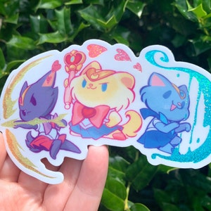 May include: A sticker featuring three cartoon characters dressed as sailor scouts. The characters are a black cat, a yellow dog, and a blue cat. The sticker has a sparkly, iridescent finish.