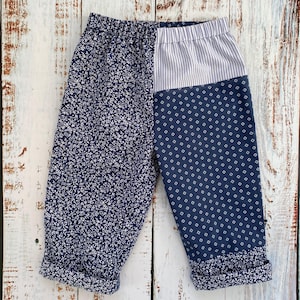 May include: Baby trousers with a patchwork design. One side has a navy blue floral print, the other a navy blue geometric pattern and a striped section. The cuffs are rolled up.