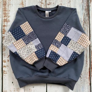 May include: A dark blue sweatshirt with patchwork sleeves. The sleeves feature a mix of patterned fabrics in navy, beige, and white. The sweatshirt has a crew neck and ribbed cuffs. The tag reads "& Love".
