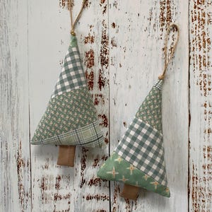 May include: Two fabric Christmas tree decorations. Each tree is made from green and white patterned fabric, with a brown wooden trunk and a jute loop. The fabric patterns include gingham, floral, and cross designs. A festive touch.