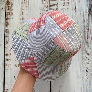 May include: A bucket hat constructed from a patchwork of fabric panels featuring various patterns including red and white stripes, blue and white pinstripes, and green plaid. The hat has a rounded crown and a downward-sloping brim.