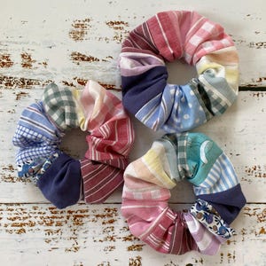May include: Three fabric scrunchies in a variety of patterns and colours, including stripes, polka dots, and floral designs. The scrunchies are arranged on a white wooden surface. The colour palette includes pink, blue, green, and purple, creating a vibrant and playful aesthetic.