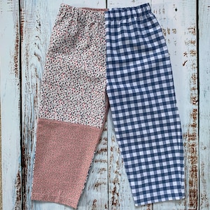 May include: Children's trousers with a distinctive design. One leg has a pink floral pattern, the other a blue and white gingham check. The trousers are made of soft fabric and have an elasticated waistband.