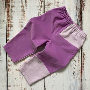 May include: Lavender and light purple baby trousers with an elasticated waistband. The trousers are made of a corduroy-like fabric and feature a colour-blocked design on the legs. They are on a weathered, light-coloured wooden surface.