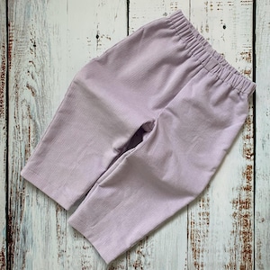 May include: Lavender corduroy trousers with an elasticated waistband. The children's trousers are made from a soft, ribbed fabric. The garment is displayed flat on a light-coloured wooden surface.