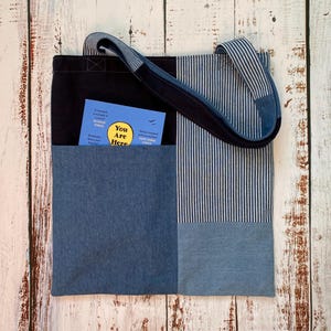 May include: A patchwork tote bag with a black top, denim pocket, and striped fabric. The bag has a denim handle and a small blue book in the pocket with the text "You Are Here".