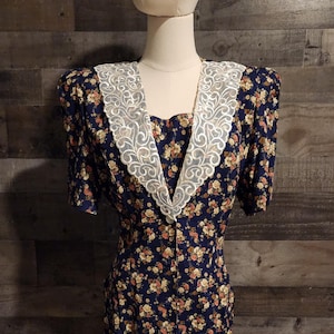 May include: A vintage navy blue dress with a floral pattern in shades of peach, yellow, and white. The dress features a white lace collar and short sleeves. It is buttoned down the front and has a fitted silhouette.