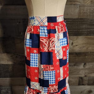 May include: A patchwork skirt in red, white, and blue, displayed on a mannequin. The skirt features a variety of patterns, including gingham, paisley, and solid blocks. A ruffled hemline adds a decorative touch to the garment.