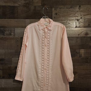 May include: A light pink, long-sleeved nightgown with a lace-trimmed collar and cuffs. The front features vertical lace panels and button closures. The nightgown is hanging on a metal hanger against a wood-paneled background.