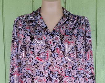 May include: A long-sleeved blouse with a dark background and a vibrant paisley floral pattern in red, white, and blue. The blouse has a collared neckline and is displayed on a mannequin.