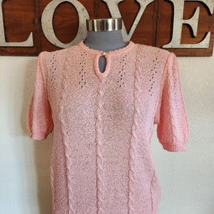 May include: A pink knitted sweater with a cable knit pattern and a keyhole neckline.