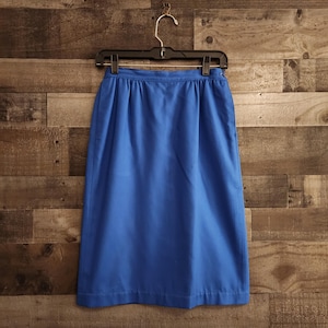 May include: A knee-length, bright blue skirt hangs on a black hanger against a wood-paneled wall. The skirt has a simple, classic design with a gathered waistline. The fabric appears to be a smooth, solid color.
