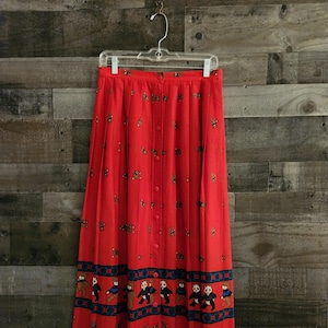 May include: A red pleated skirt with a button-up front and a pattern of brown bears and pandas. The skirt has a blue and black trim at the bottom.