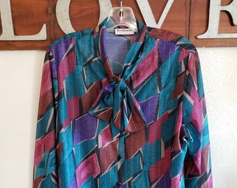 May include: A teal, purple, pink, and brown geometric patterned blouse with a bow tie. The blouse has a button-up front and long sleeves.