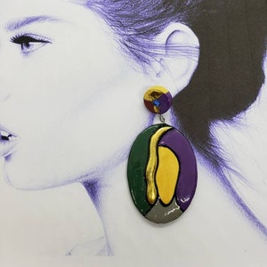 May include: A pair of colorful, abstract oval-shaped earrings. The earrings are made of clay and feature a green, yellow, purple, and gray color scheme. The earrings are hanging from a silver hook.