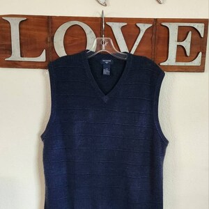 May include: A navy blue sleeveless sweater vest with a V-neck and a subtle textured pattern. The vest is hanging on a hanger with the brand label "Dockers" visible.