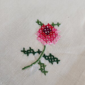 Pu&ograve; includere: Primo piano di un ricamo floreale cucito a mano. Il disegno presenta una rosa rosa e rossa con foglie e stelo verdi su un tessuto color crema. Il ricamo &egrave; realizzato a punto croce.