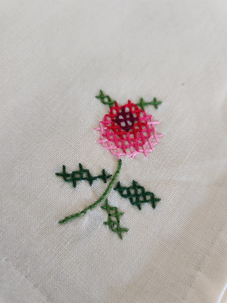 Pu&ograve; includere: Primo piano di un ricamo floreale cucito a mano. Il disegno presenta una rosa rosa e rossa con foglie e stelo verdi su un tessuto color crema. Il ricamo &egrave; realizzato a punto croce.