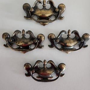 May include: Four antique brass drawer pulls with ornate, curved designs. Each pull features a central decorative element and a curved handle. The brass has a slightly aged appearance, adding to the vintage aesthetic. Perfect for furniture restoration projects.