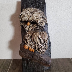 May include: A brown and white owl figurine perched on a branch, mounted on a dark wood plaque. The owl has a textured, feathery appearance and a small orange beak.
