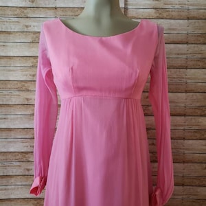 May include: A long-sleeved pink dress featuring a textured fabric, a rounded neckline, and a high waistline. The sleeves are sheer and end in buttoned cuffs. The bodice is fitted, while the skirt flows downward from the empire waistline.