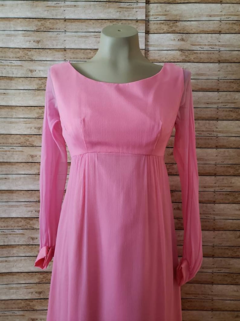 May include: A long-sleeved pink dress featuring a textured fabric, a rounded neckline, and a high waistline. The sleeves are sheer and end in buttoned cuffs. The bodice is fitted, while the skirt flows downward from the empire waistline.