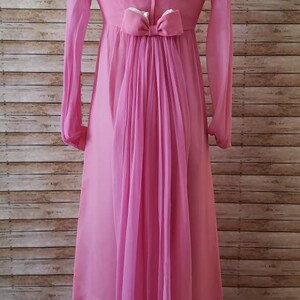 May include: A floor-length pink gown featuring long, sheer sleeves and a V-neckline. The back of the dress includes a central vertical seam, a small bow detail at the waist, and a flowing, pleated sheer panel that extends down to the hemline.