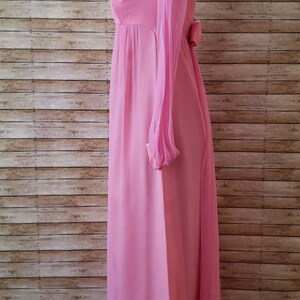 May include: A floor-length, solid pink dress featuring long, sheer sleeves and a high neckline. The garment has a fitted bodice that transitions into a flowing skirt, with a small decorative bow detail visible at the waistline on the side profile.