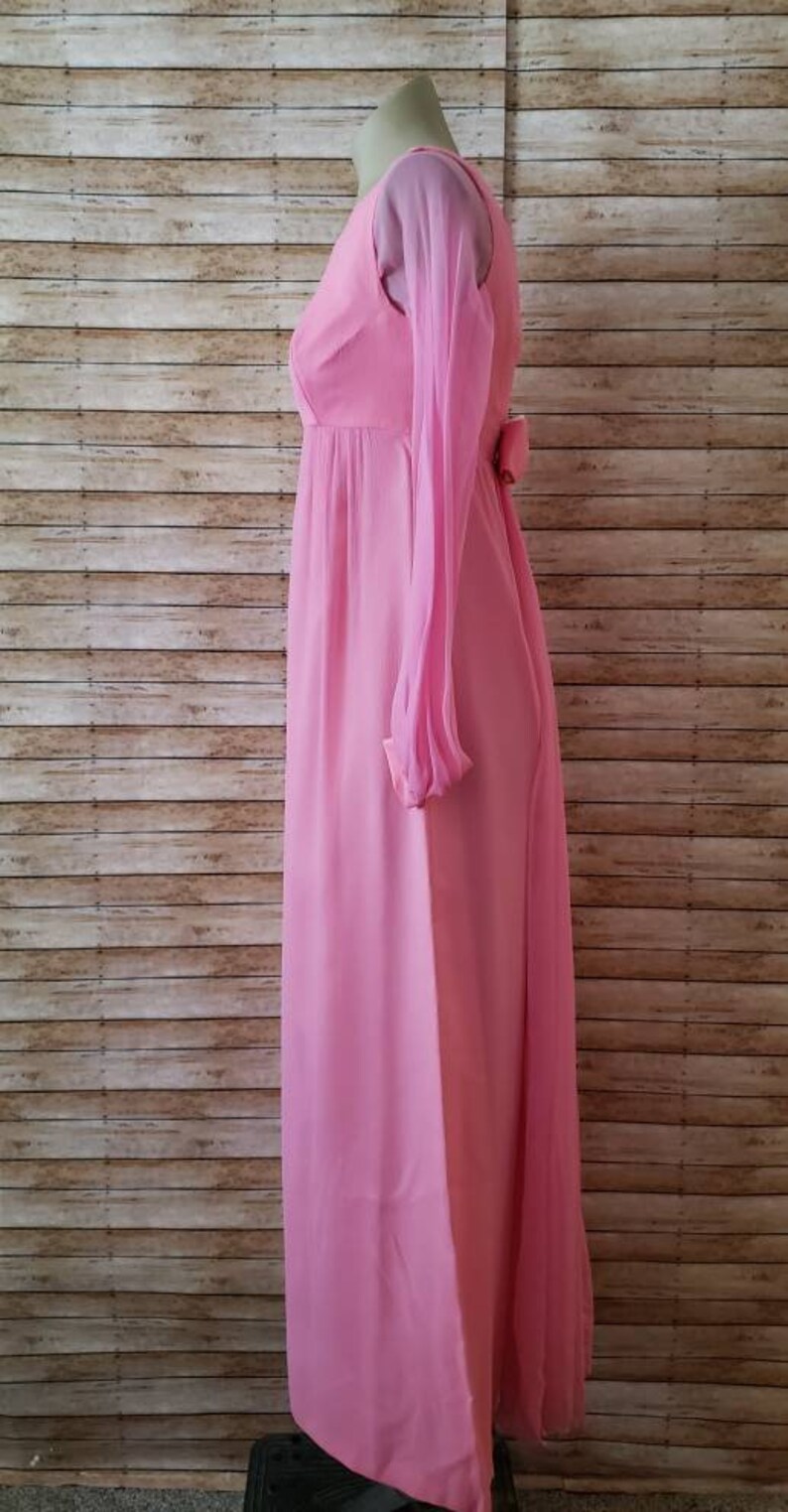 May include: A floor-length, solid pink dress featuring long, sheer sleeves and a high neckline. The garment has a fitted bodice that transitions into a flowing skirt, with a small decorative bow detail visible at the waistline on the side profile.