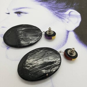 May include: A pair of black oval-shaped earrings with a smaller round accent piece. The accent piece is a layered design of black, gold, and purple.
