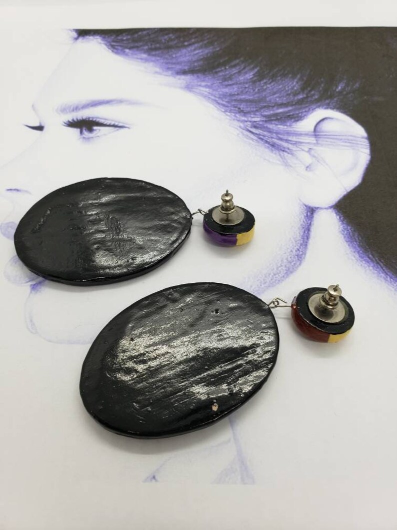 May include: A pair of black oval-shaped earrings with a smaller round accent piece. The accent piece is a layered design of black, gold, and purple.