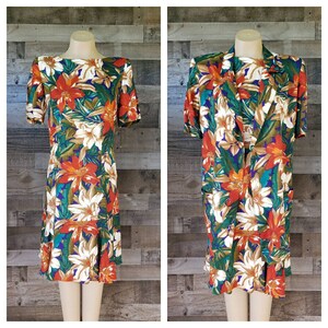 May include: Two-piece floral outfit featuring a dress and a matching jacket. The dress has a boat neck and short sleeves, with a ruffled hem. The jacket has a notched collar and short sleeves. Both pieces are adorned with a vibrant floral pattern in red, orange, white, green, and blue.